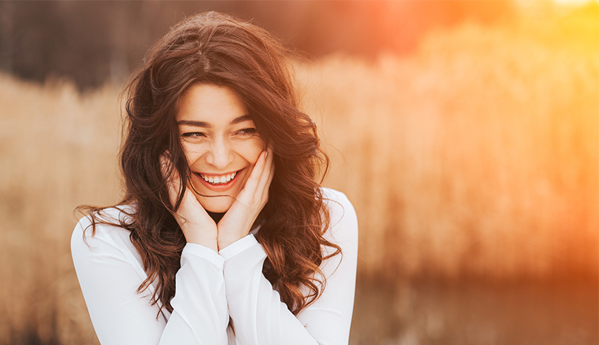 A smiling woman stands outside with her hands placed on either side of her face